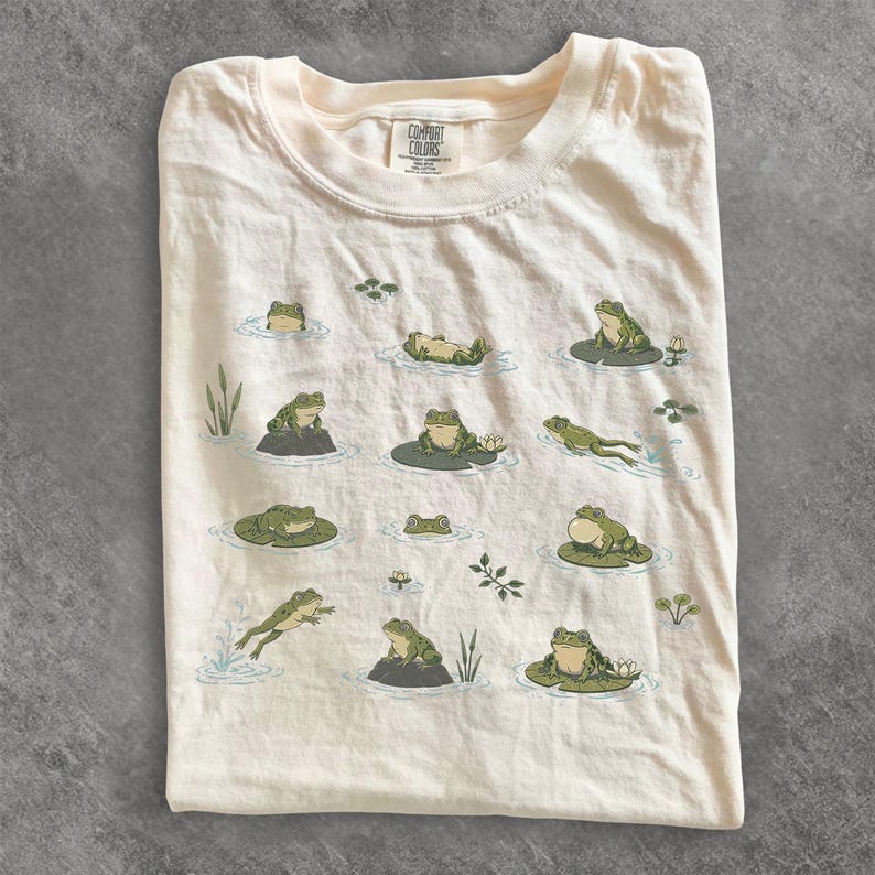 May include: A cream-colored t-shirt featuring a repeating graphic print of various green frogs in different poses, including swimming, sitting on lily pads, and resting on rocks. The shirt has a crew neckline and a Comfort Colors brand label.
