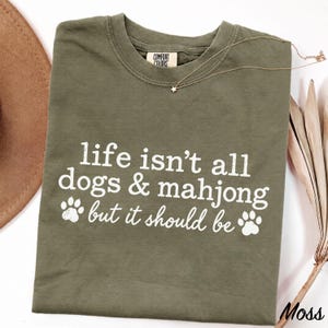 May include: An olive green crew neck t-shirt with white screen-printed text that reads, life isn't all dogs & mahjong but it should be, accompanied by two white paw print graphics. The shirt features a Comfort Colors brand neck label.