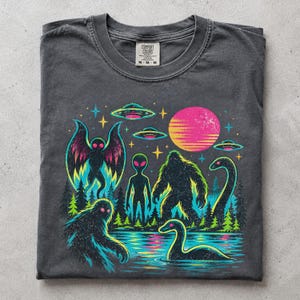 May include: A charcoal gray t-shirt featuring a vibrant graphic print of cryptids and extraterrestrial elements. The design includes a moth-like creature, an alien, a sasquatch, and a sea serpent near a lake, with UFOs and a bright pink sun above.
