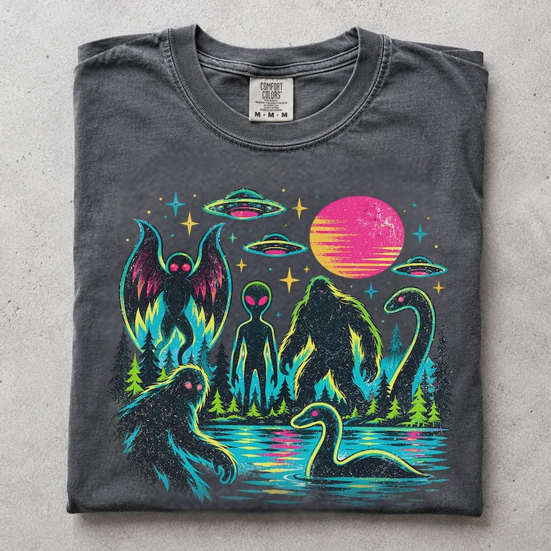 May include: A charcoal gray t-shirt featuring a vibrant graphic print of cryptids and extraterrestrial elements. The design includes a moth-like creature, an alien, a sasquatch, and a sea serpent near a lake, with UFOs and a bright pink sun above.