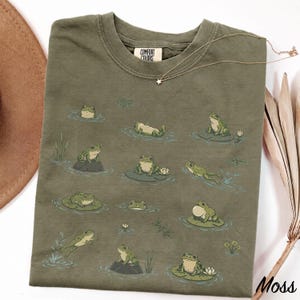 May include: An olive green crew neck t-shirt featuring a repeating graphic print of various green frogs in different poses, including swimming, sitting on lily pads, and jumping. The shirt has a Comfort Colors brand label at the inner neckline.