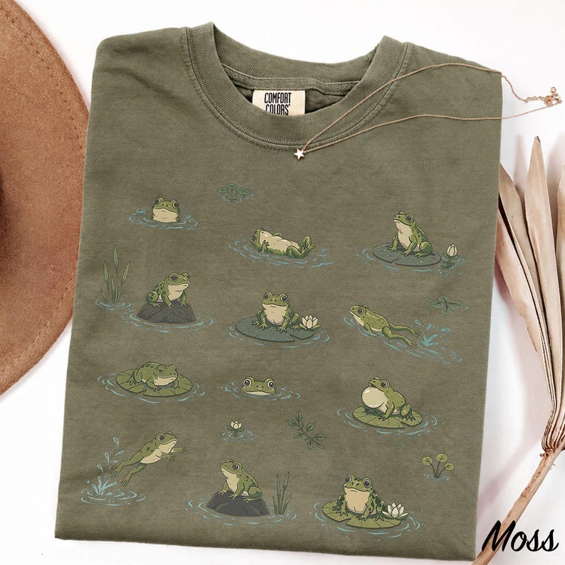 May include: An olive green crew neck t-shirt featuring a repeating graphic print of various green frogs in different poses, including swimming, sitting on lily pads, and jumping. The shirt has a Comfort Colors brand label at the inner neckline.