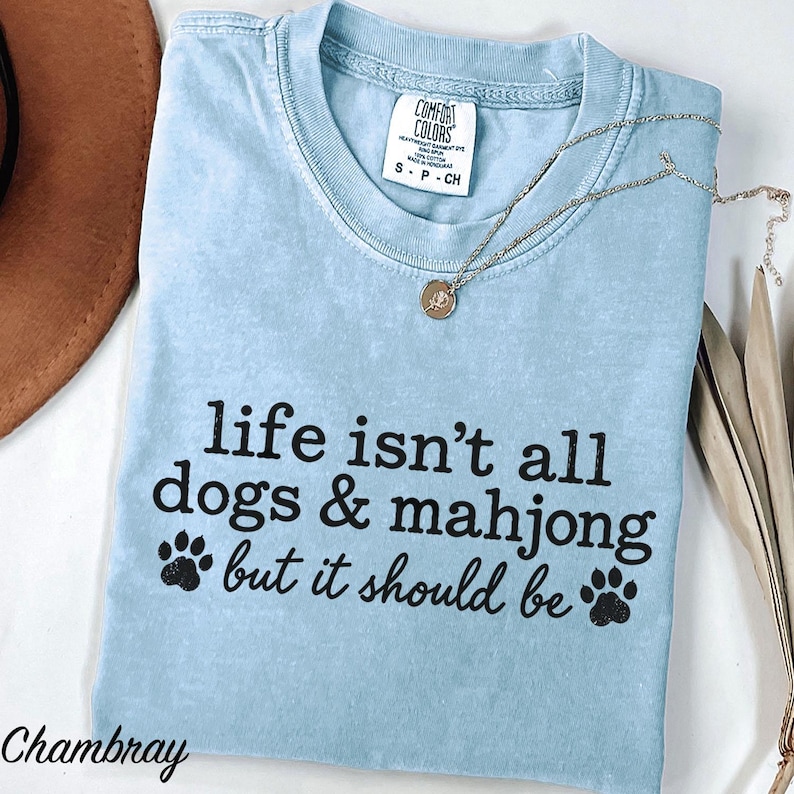 May include: A light blue t-shirt with a crew neckline and a Comfort Colors label at the collar. The shirt features black text that reads, "life isn't all dogs & mahjong but it should be," accompanied by two black paw print graphics on the front.