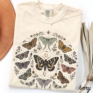 Op de afbeelding: Een ivoorkleurig t-shirt met een cirkelvormige grafische print van diverse motten en vlinders in bruin-, groen-, blauw- en rozetinten, gerangschikt rond een grote centrale mot met maansikkels en kleine bloemtakjes.