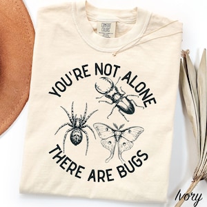 Op de afbeelding: Een ivoorkleurig t-shirt met ronde hals en een zwarte grafische print van een spin, een kever en een mot. De tekst "YOU'RE NOT ALONE THERE ARE BUGS" is in een cirkel rond de insecten geplaatst. Het shirt heeft een Comfort Colors merklabel.
