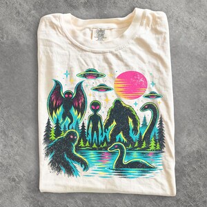 May include: A cream-colored t-shirt featuring a graphic print of a mothman, an alien, a bigfoot, and a sea creature in a forest setting with UFOs and a pink sun. The shirt has a Comfort Colors label at the neck and is made of 100% cotton.