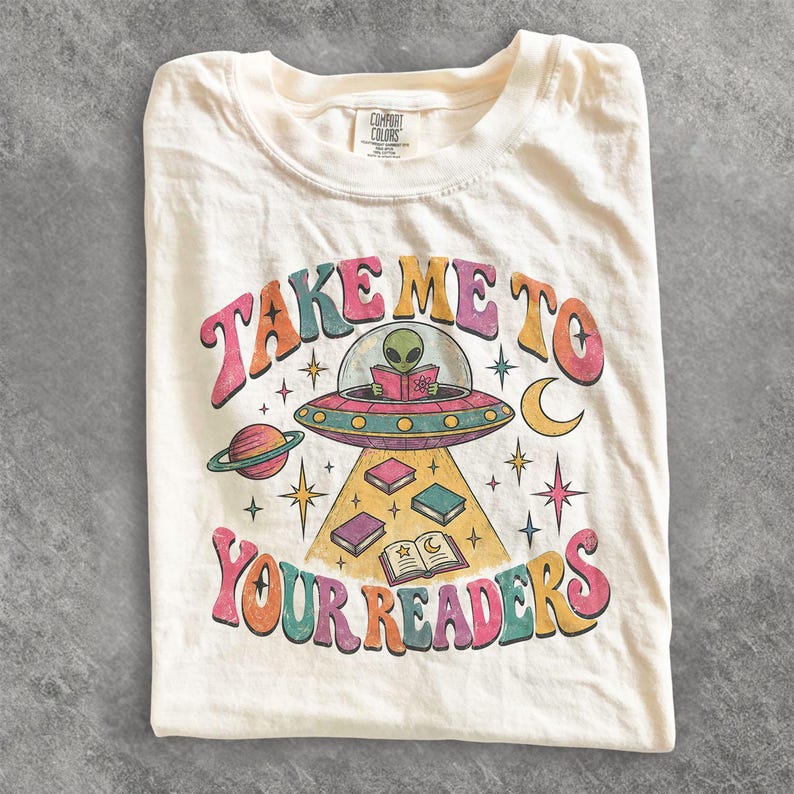 May include: A cream-coloured t-shirt featuring a graphic of a green alien in a flying saucer reading a book. The text reads "TAKE ME TO YOUR READERS" in colourful, curved lettering. The design includes planets, stars, and floating books.
