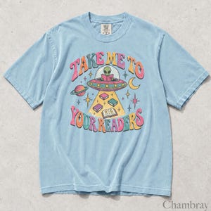 May include: A light blue short-sleeve t-shirt featuring a graphic of an alien in a flying saucer reading a book. The text "TAKE ME TO YOUR READERS" is printed in a colorful, wavy font. The design includes planets, stars, and floating books.