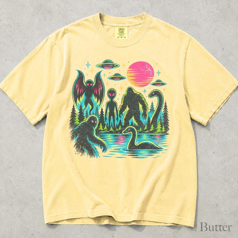 May include: A pale yellow short-sleeve t-shirt featuring a graphic print of cryptids including Bigfoot, Mothman, and the Loch Ness Monster, alongside an alien, UFOs, a forest, and a pink sun. The design uses vibrant neon colors against a dark background.
