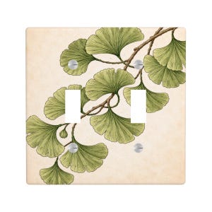 Peut inclure: Un cache-interrupteur double avec une illustration botanique de feuilles et de branches de ginkgo en vert et marron. Le motif est posé sur un fond crème, avec des vis argentées fixant le cache.