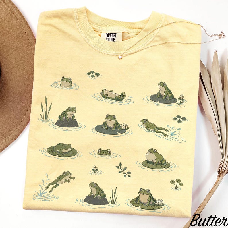 May include: A pale yellow t-shirt featuring a repeating graphic print of green frogs in various poses, including swimming, sitting on lily pads, and resting on rocks. The shirt has a crew neckline and a Comfort Colors brand label at the collar.