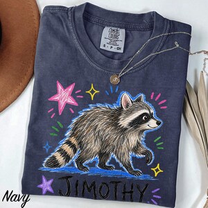 May include: A navy blue short-sleeve t-shirt featuring a graphic of a raccoon walking, surrounded by colorful hand-drawn style stars and dashes. The text "JIMOTHY" is printed in black below the raccoon. The shirt has a Comfort Colors brand label.