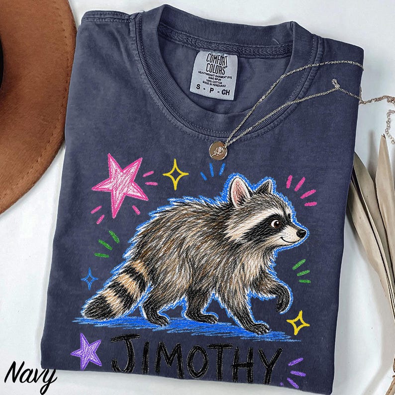 May include: A navy blue short-sleeve t-shirt featuring a graphic of a raccoon walking, surrounded by colorful hand-drawn style stars and dashes. The text "JIMOTHY" is printed in black below the raccoon. The shirt has a Comfort Colors brand label.