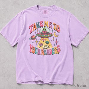 May include: A light purple short-sleeve t-shirt featuring a graphic of an alien in a flying saucer reading a book. The text "TAKE ME TO YOUR READERS" is printed in a colorful, wavy font. The design includes stars, a planet, and books.