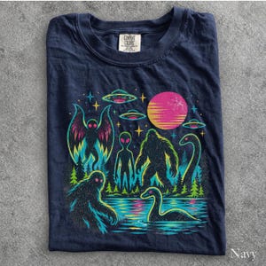 May include: A navy blue t-shirt featuring a colorful graphic print of a moth-like creature, an alien, a bigfoot, and a long-necked sea monster in a forest setting under a pink sun and UFOs. The shirt has a Comfort Colors brand label at the neck.