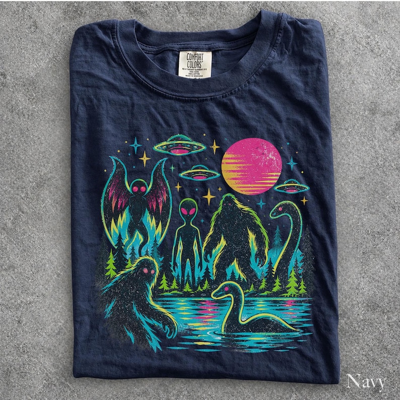 May include: A navy blue t-shirt featuring a colorful graphic print of a moth-like creature, an alien, a bigfoot, and a long-necked sea monster in a forest setting under a pink sun and UFOs. The shirt has a Comfort Colors brand label at the neck.