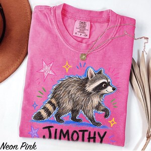 May include: A neon pink short-sleeved t-shirt featuring a graphic illustration of a raccoon walking. The design includes colorful hand-drawn style stars and the name JIMOTHY printed in bold black letters below the raccoon. The shirt has a crew neckline.