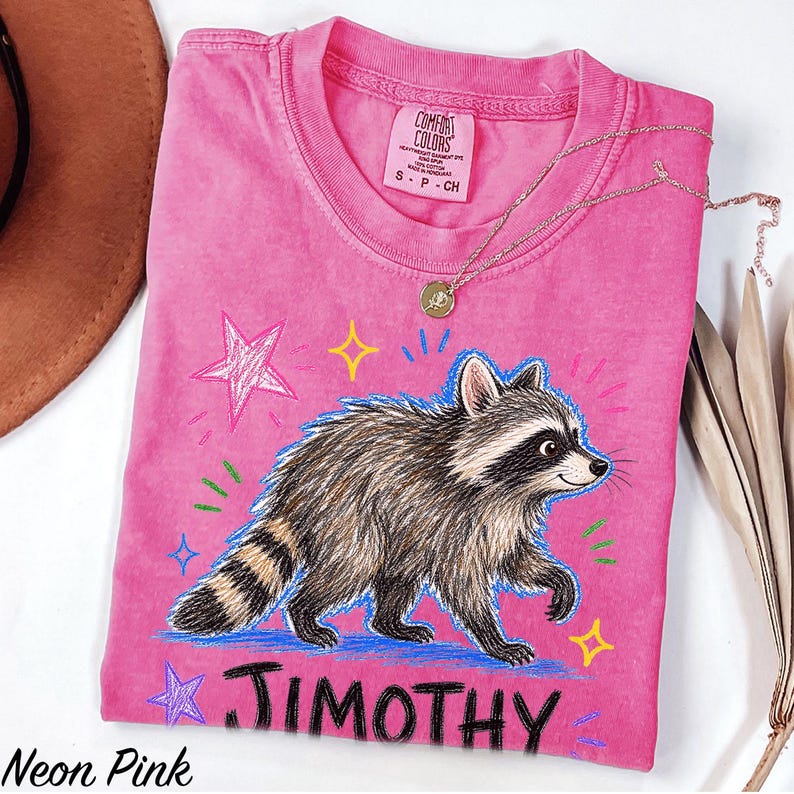 May include: A neon pink short-sleeved t-shirt featuring a graphic illustration of a raccoon walking. The design includes colorful hand-drawn style stars and the name JIMOTHY printed in bold black letters below the raccoon. The shirt has a crew neckline.
