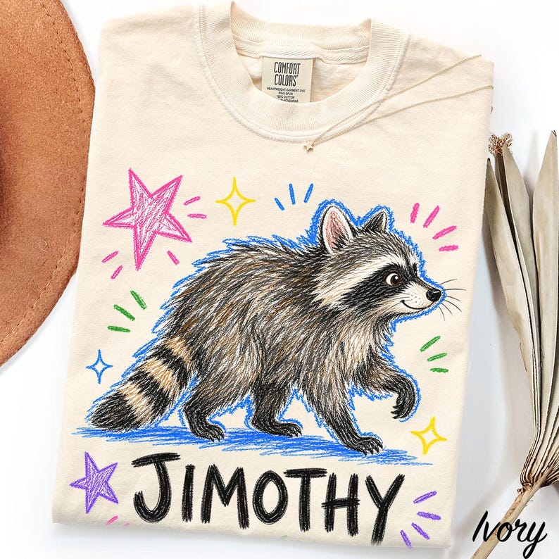 May include: An ivory t-shirt featuring a graphic of a raccoon with a black and white mask and striped tail. The design includes colorful hand-drawn style stars and the name JIMOTHY in bold black lettering. The shirt has a Comfort Colors brand label.