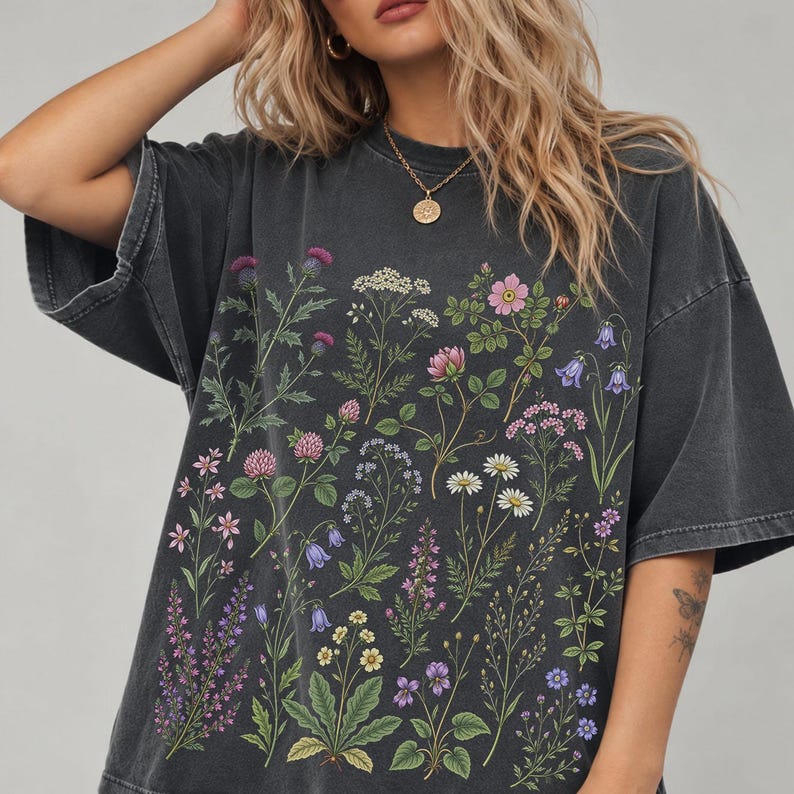 May include: A charcoal gray short-sleeve t-shirt featuring a detailed botanical print of various wildflowers in shades of pink, purple, blue, white, and green. The shirt has a relaxed fit with a crew neckline and a faded, washed-out fabric texture.