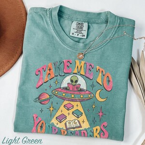 May include: A light green t-shirt featuring a graphic of an alien in a flying saucer reading a book. The text reads "TAKE ME TO YOUR READERS" in colorful, retro-style lettering. The design includes planets, stars, and floating books.