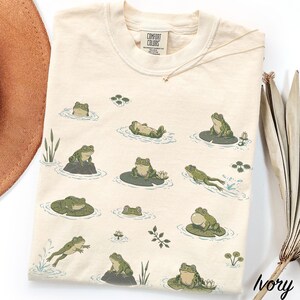 May include: An ivory t-shirt featuring a repeating pattern of green frogs in various poses, including swimming, sitting on lily pads, and resting on rocks. The shirt has a crew neckline and a Comfort Colors brand label printed at the inner collar.