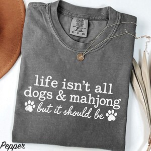 May include: A charcoal gray short-sleeve t-shirt with a crew neckline and white screen-printed text that reads, life isn't all dogs & mahjong but it should be, accompanied by two white paw print graphics on the front of the garment.