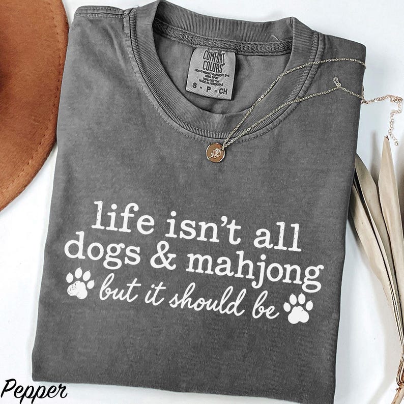 May include: A charcoal gray short-sleeve t-shirt with a crew neckline and white screen-printed text that reads, life isn't all dogs & mahjong but it should be, accompanied by two white paw print graphics on the front of the garment.