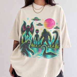 May include: A cream-colored short-sleeve t-shirt featuring a vibrant graphic print of a forest scene with a moth-like creature, an alien, a Bigfoot, and a Loch Ness monster, alongside flying saucers, stars, and a pink sun in the background.