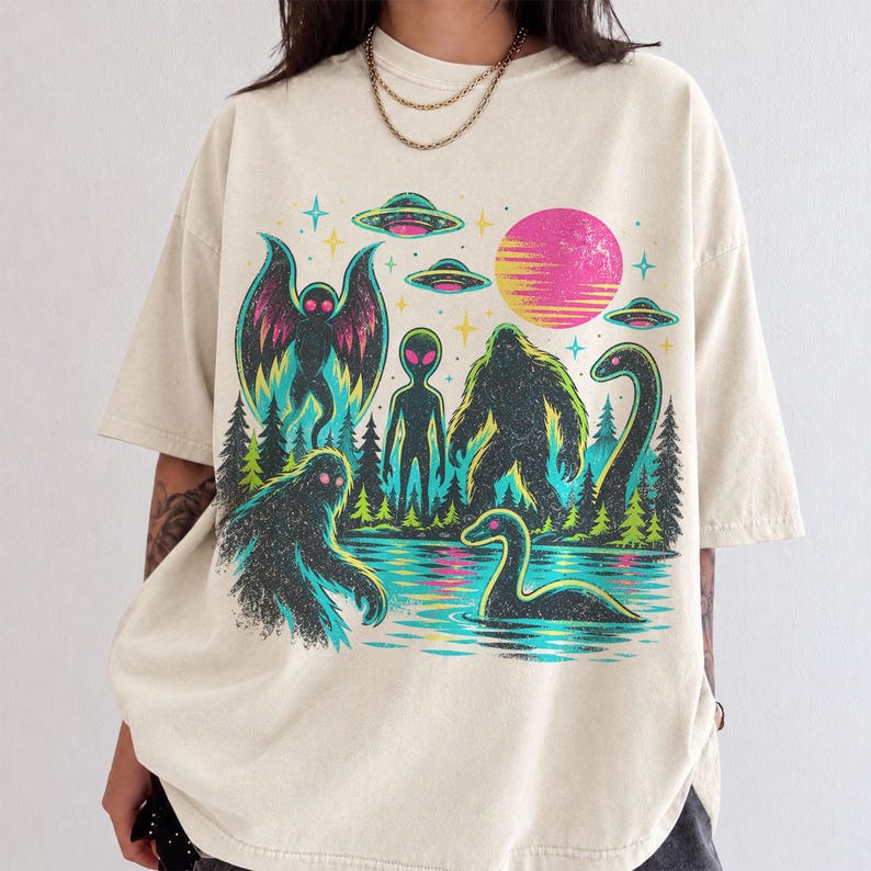 May include: A cream-colored short-sleeve t-shirt featuring a vibrant graphic print of a forest scene with a moth-like creature, an alien, a Bigfoot, and a Loch Ness monster, alongside flying saucers, stars, and a pink sun in the background.
