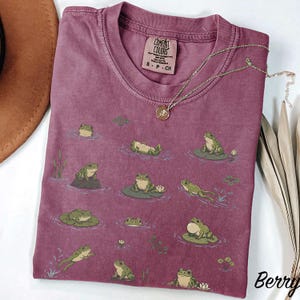 May include: A berry-colored t-shirt featuring a repeating graphic print of green frogs in various poses on lily pads and in water. The shirt has a crew neckline and a Comfort Colors brand label on the inside collar. A gold-toned necklace is placed on top.