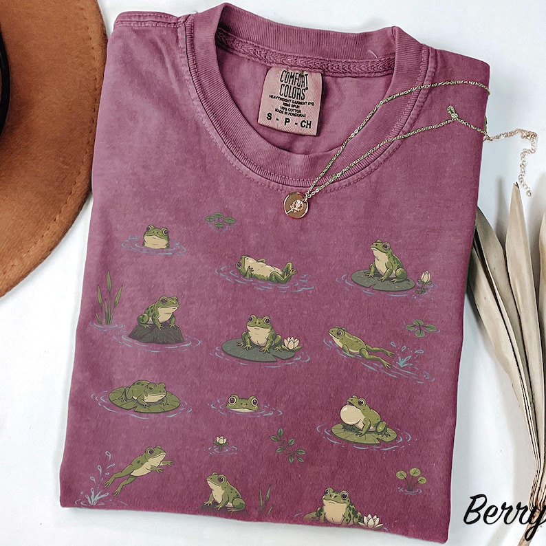 May include: A berry-colored t-shirt featuring a repeating graphic print of green frogs in various poses on lily pads and in water. The shirt has a crew neckline and a Comfort Colors brand label on the inside collar. A gold-toned necklace is placed on top.