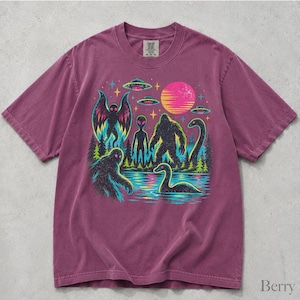May include: A berry-colored short-sleeve t-shirt featuring a graphic print of a mothman, an alien, a bigfoot, and a loch ness monster near a lake. The design includes flying saucers, a bright pink sun, stars, and pine trees in neon colors.