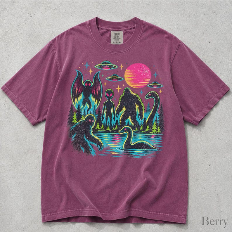 May include: A berry-colored short-sleeve t-shirt featuring a graphic print of a mothman, an alien, a bigfoot, and a loch ness monster near a lake. The design includes flying saucers, a bright pink sun, stars, and pine trees in neon colors.