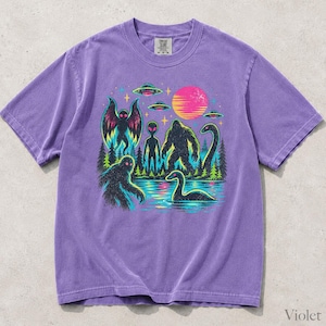 May include: A purple short-sleeve t-shirt featuring a graphic print of cryptids including Mothman, Bigfoot, and the Loch Ness Monster, alongside an alien, UFOs, a forest, and a pink sun. The shirt has a crew neckline and a tag at the collar.