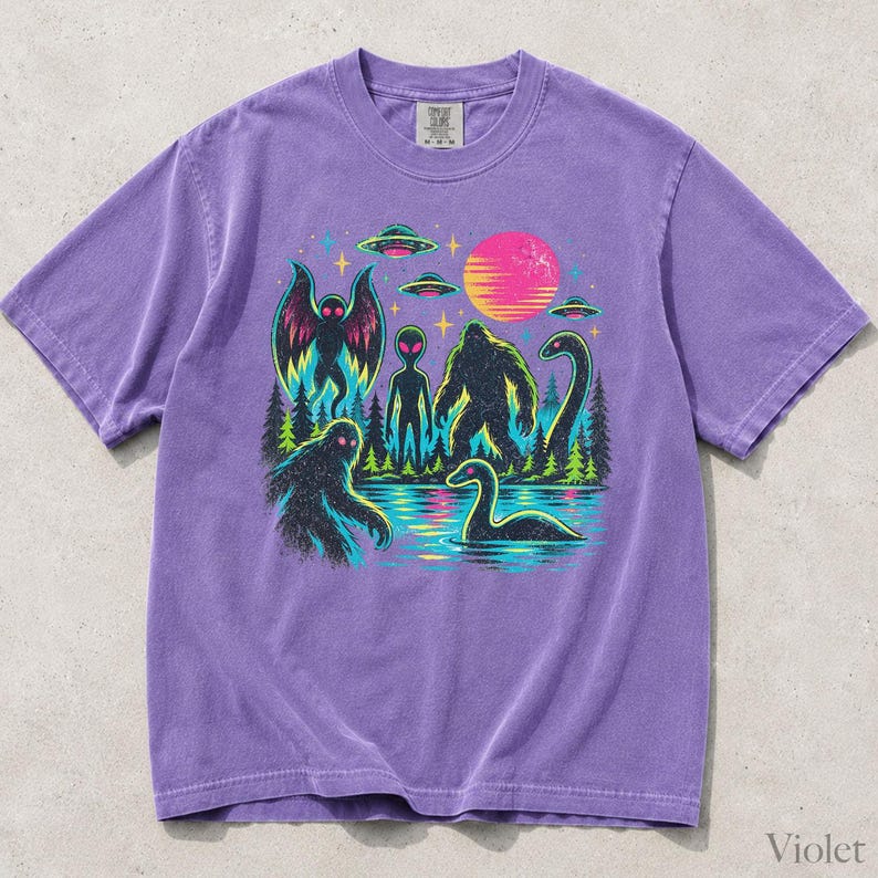 May include: A purple short-sleeve t-shirt featuring a graphic print of cryptids including Mothman, Bigfoot, and the Loch Ness Monster, alongside an alien, UFOs, a forest, and a pink sun. The shirt has a crew neckline and a tag at the collar.