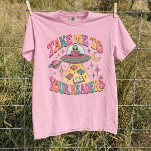 May include: A light pink short-sleeve t-shirt featuring a graphic of a green alien in a flying saucer reading a book. The text reads "TAKE ME TO YOUR READERS" in colorful, retro-style lettering, surrounded by floating books, stars, and a planet.