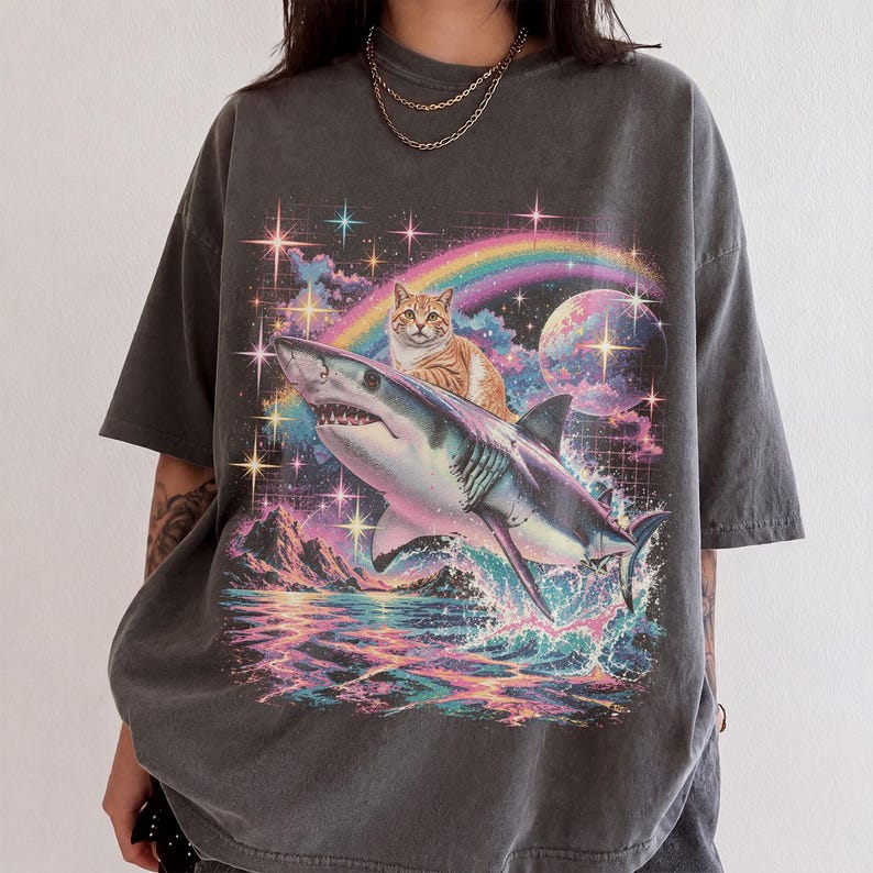 May include: A charcoal gray t-shirt featuring a graphic print of a tabby cat riding a shark over a colorful ocean scene. The design includes a rainbow, a large planet, and sparkling stars against a dark, grid-patterned cosmic background.