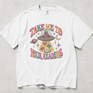 May include: A white short-sleeve t-shirt featuring a colorful graphic of an alien in a flying saucer reading a book. The text "TAKE ME TO YOUR READERS" surrounds the illustration, which includes floating books, a planet, a crescent moon, and stars.
