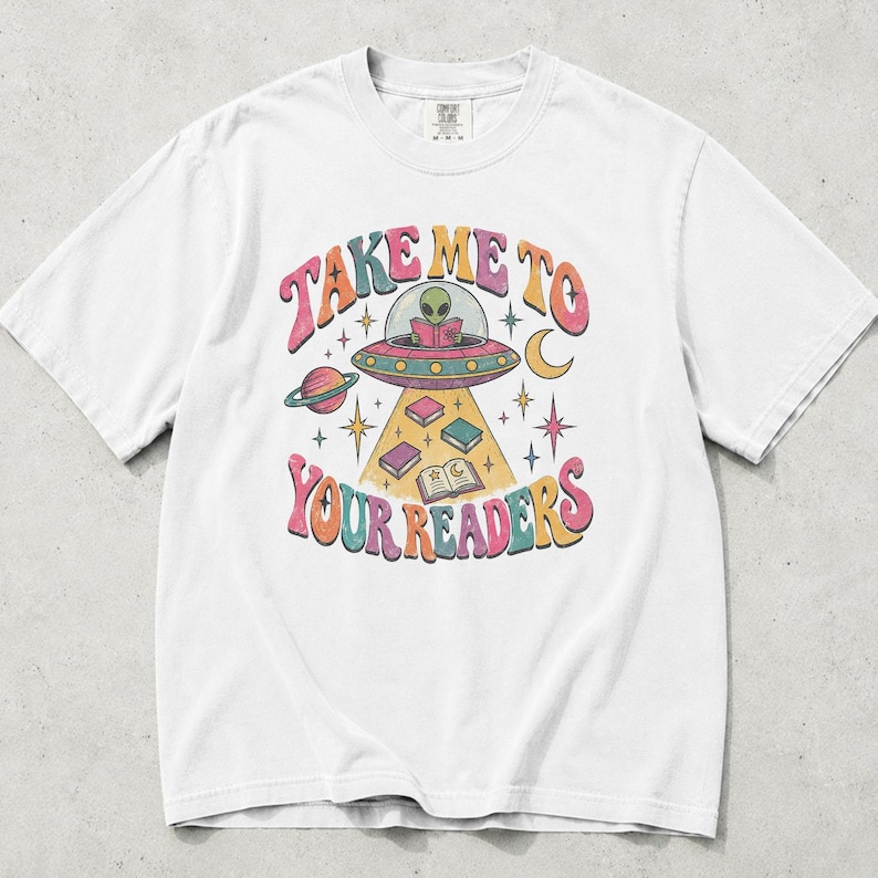 May include: A white short-sleeve t-shirt featuring a colorful graphic of an alien in a flying saucer reading a book. The text "TAKE ME TO YOUR READERS" surrounds the illustration, which includes floating books, a planet, a crescent moon, and stars.