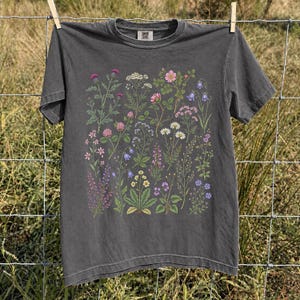 May include: A charcoal gray short-sleeve t-shirt featuring a detailed botanical print of various wildflowers and plants in shades of pink, purple, white, yellow, and green. The shirt has a crew neckline and a visible brand label at the inner collar.