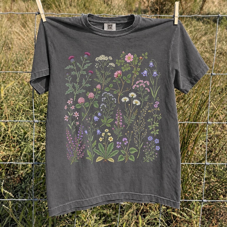May include: A charcoal gray short-sleeve t-shirt featuring a detailed botanical print of various wildflowers and plants in shades of pink, purple, white, yellow, and green. The shirt has a crew neckline and a visible brand label at the inner collar.