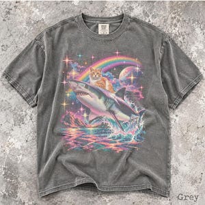 May include: A charcoal grey short-sleeve t-shirt featuring a graphic print of an orange tabby cat riding a shark over a colorful ocean scene. The design includes a rainbow, a planet, and sparkling stars. The shirt has a crew neckline and a label.