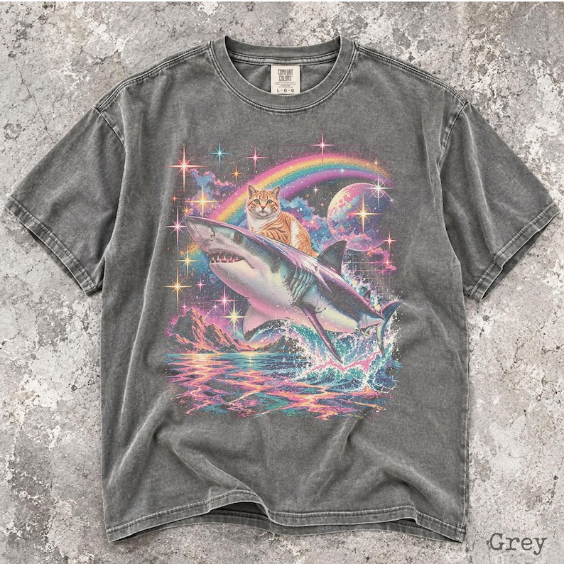 May include: A charcoal grey short-sleeve t-shirt featuring a graphic print of an orange tabby cat riding a shark over a colorful ocean scene. The design includes a rainbow, a planet, and sparkling stars. The shirt has a crew neckline and a label.