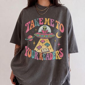 May include: A charcoal gray short-sleeve t-shirt featuring a graphic print of an alien in a flying saucer reading a book. The text reads "TAKE ME TO YOUR READERS" in colorful, retro-style lettering, surrounded by planets, stars, and floating books.