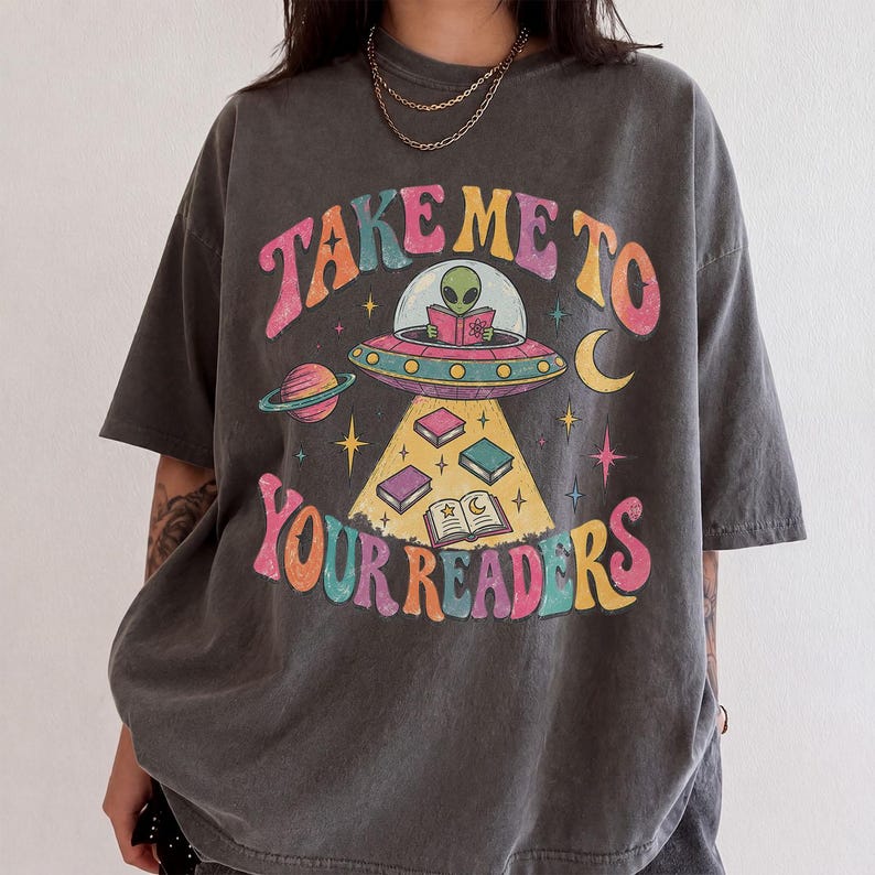 May include: A charcoal gray short-sleeve t-shirt featuring a graphic print of an alien in a flying saucer reading a book. The text reads "TAKE ME TO YOUR READERS" in colorful, retro-style lettering, surrounded by planets, stars, and floating books.