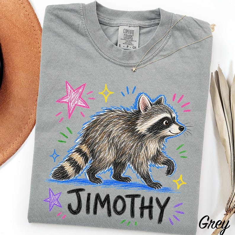 May include: A grey t-shirt featuring a graphic of a raccoon walking, surrounded by colorful hand-drawn style stars and sparkles. The name JIMOTHY is printed in large black capital letters below the raccoon. The shirt has a crew neck collar.
