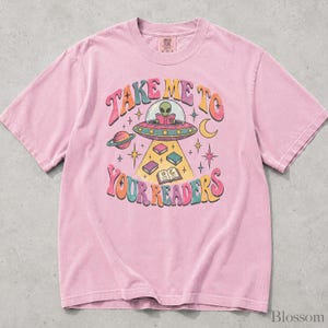May include: A light pink short-sleeve t-shirt featuring a graphic of an alien in a flying saucer reading a book. The text reads "TAKE ME TO YOUR READERS" in colorful, curved lettering, surrounded by stars, a planet, and floating books.