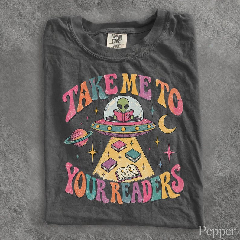 May include: A charcoal gray t-shirt featuring a graphic of an alien in a flying saucer reading a book. The text reads "TAKE ME TO YOUR READERS" in colorful, wavy letters. The design includes a planet, stars, a crescent moon, and books.