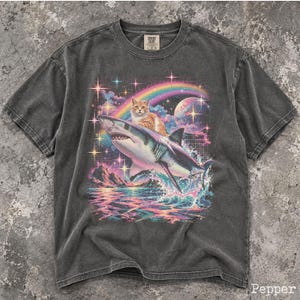 May include: A charcoal gray short-sleeve t-shirt featuring a graphic print of an orange tabby cat riding a shark. The design includes a colorful rainbow, a large planet, sparkling stars, and ocean waves. The shirt has a crew neck and a label.
